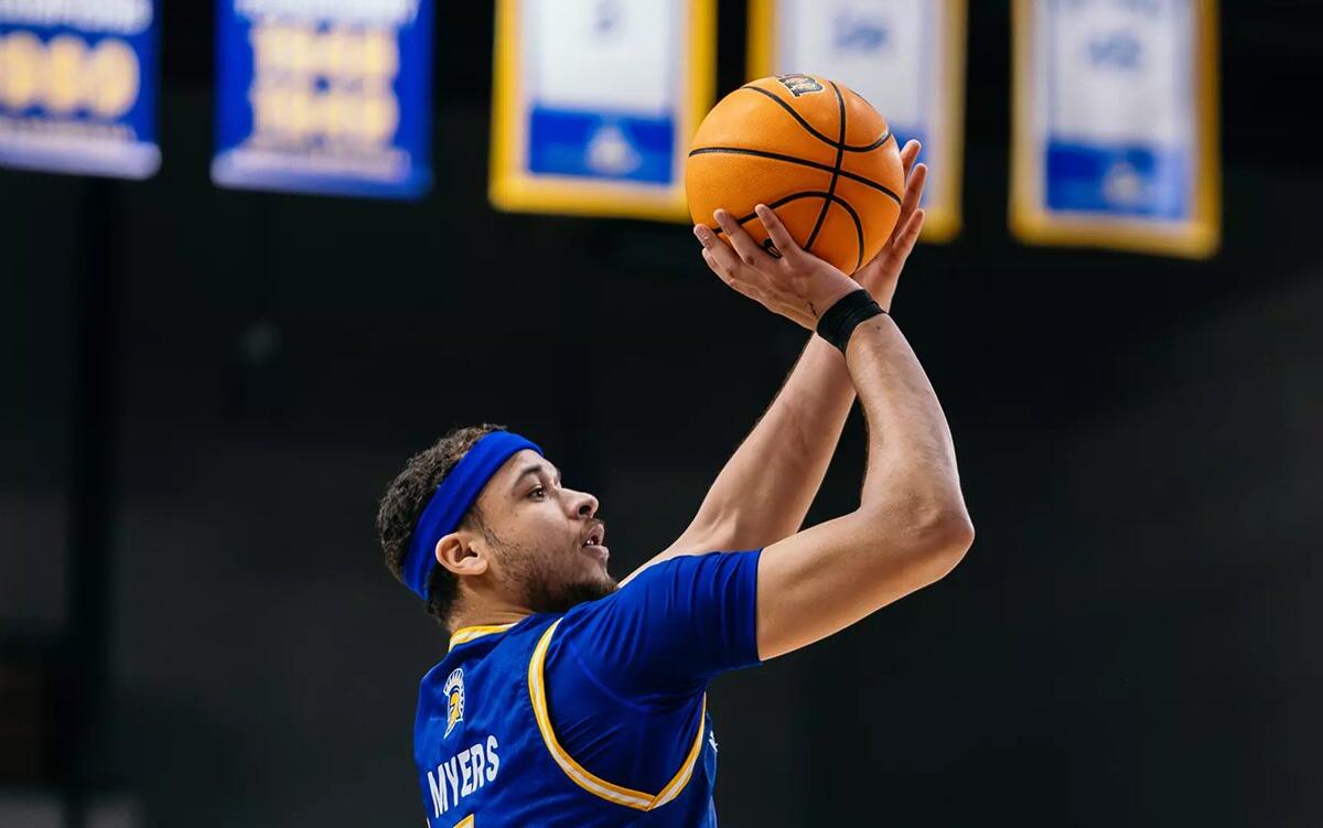 Basketball player in a blue jersey (name Myers on back) jumping to shoot a ball above his head.