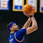 Basketball player in a blue jersey (name Myers on back) jumping to shoot a ball above his head.