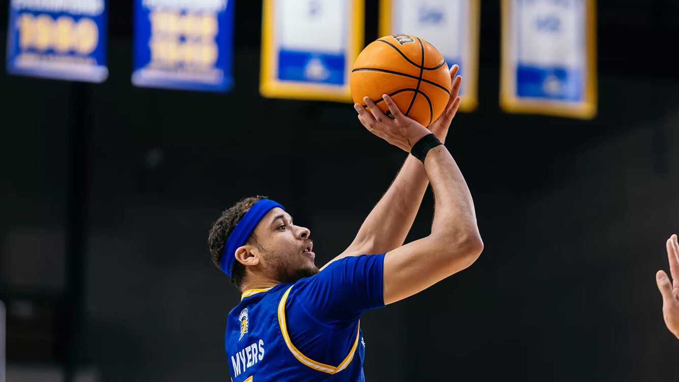 Basketball player in a blue jersey (name Myers on back) jumping to shoot a ball above his head.
