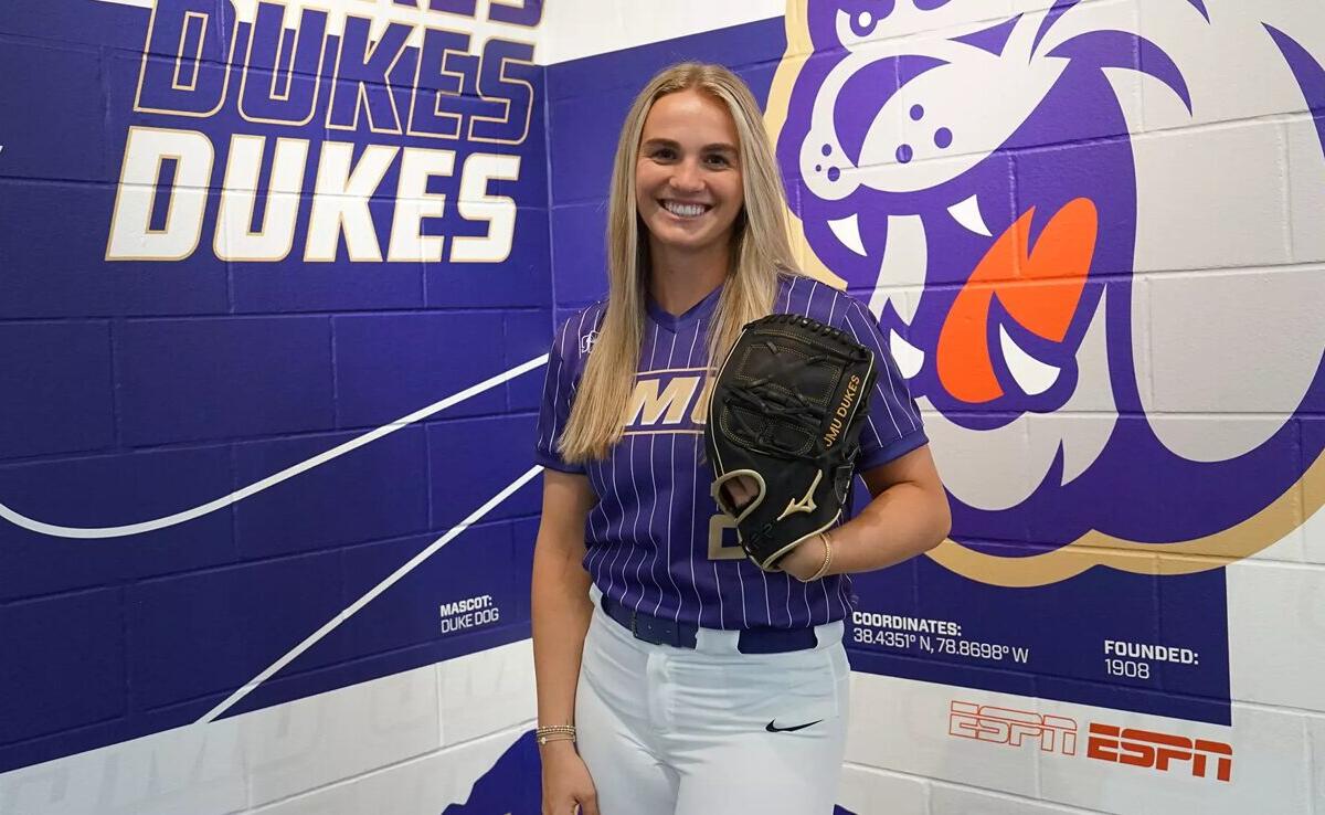 Smiling woman in a purple Duke Dukes softball uniform holds a glove, standing in front of a purple team mural.