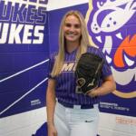 Smiling woman in a purple Duke Dukes softball uniform holds a glove, standing in front of a purple team mural.