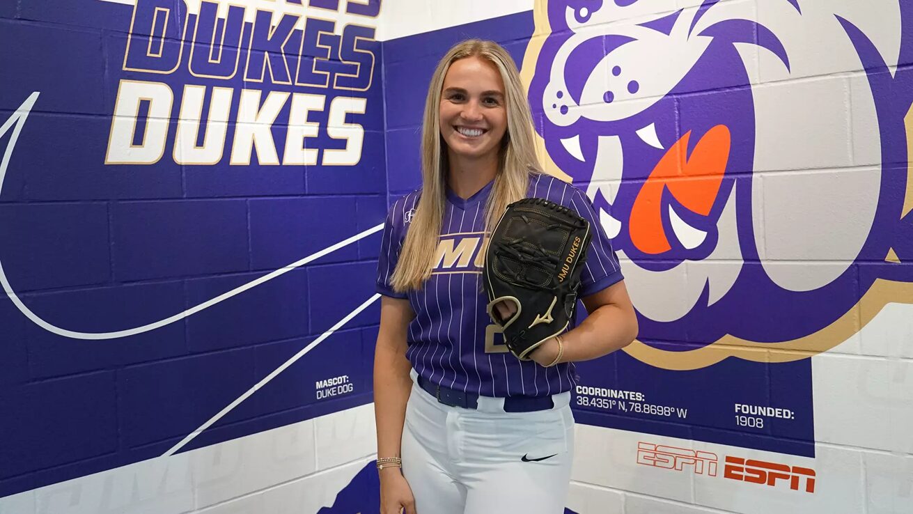 Smiling woman in a purple Duke Dukes softball uniform holds a glove, standing in front of a purple team mural.