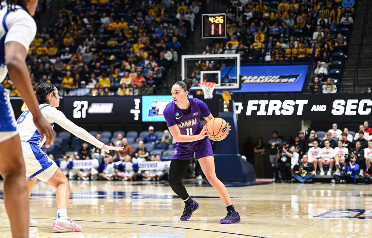 JMU player (#11) in purple dribbles the ball as a white-blue defender reaches in, in a crowded basketball arena with a visible scoreboard and crowd.