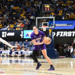 JMU player (#11) in purple dribbles the ball as a white-blue defender reaches in, in a crowded basketball arena with a visible scoreboard and crowd.