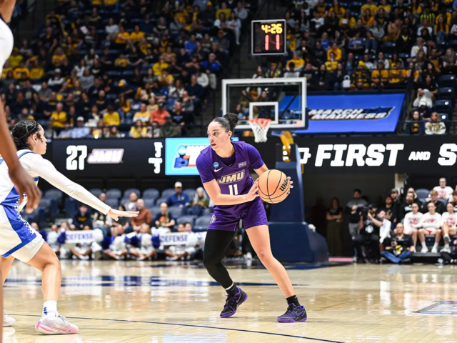 JMU player (#11) in purple dribbles the ball as a white-blue defender reaches in, in a crowded basketball arena with a visible scoreboard and crowd.