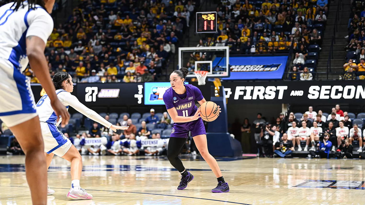 JMU player (#11) in purple dribbles the ball as a white-blue defender reaches in, in a crowded basketball arena with a visible scoreboard and crowd.