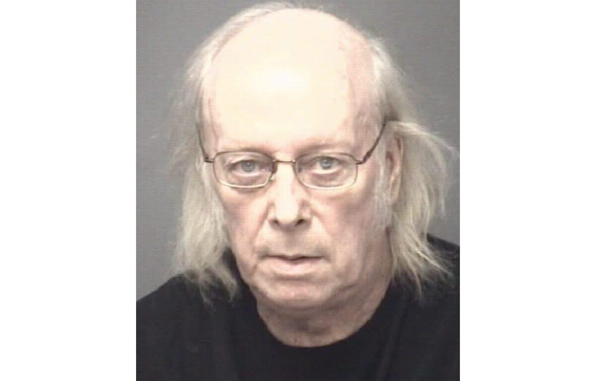 Mugshot of an older man with thin glasses, balding head and shoulder‑length gray hair, wearing a black shirt.