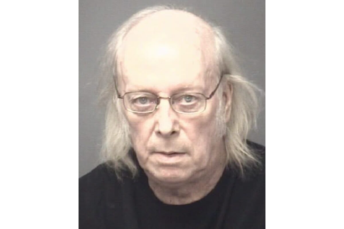 Mugshot of an older man with thin glasses, balding head and shoulder‑length gray hair, wearing a black shirt.