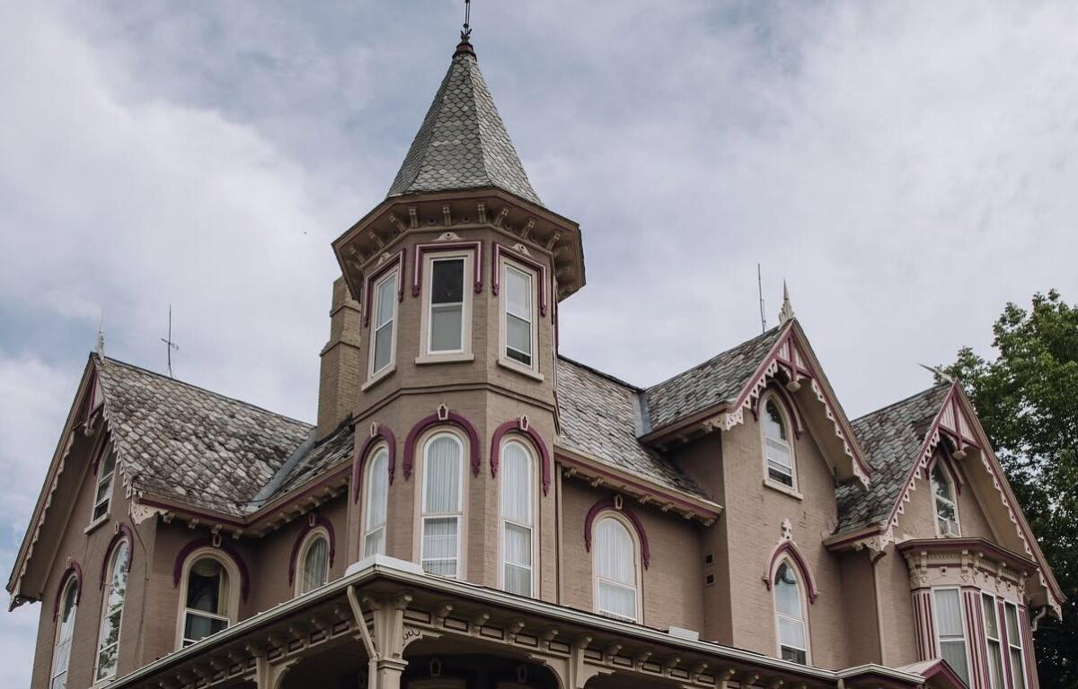 Victorian-style house with a turret, beige walls, and pink decorative trim under a cloudy sky.