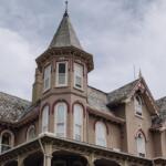 Victorian-style house with a turret, beige walls, and pink decorative trim under a cloudy sky.