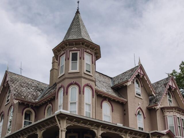 Victorian-style house with a turret, beige walls, and pink decorative trim under a cloudy sky.