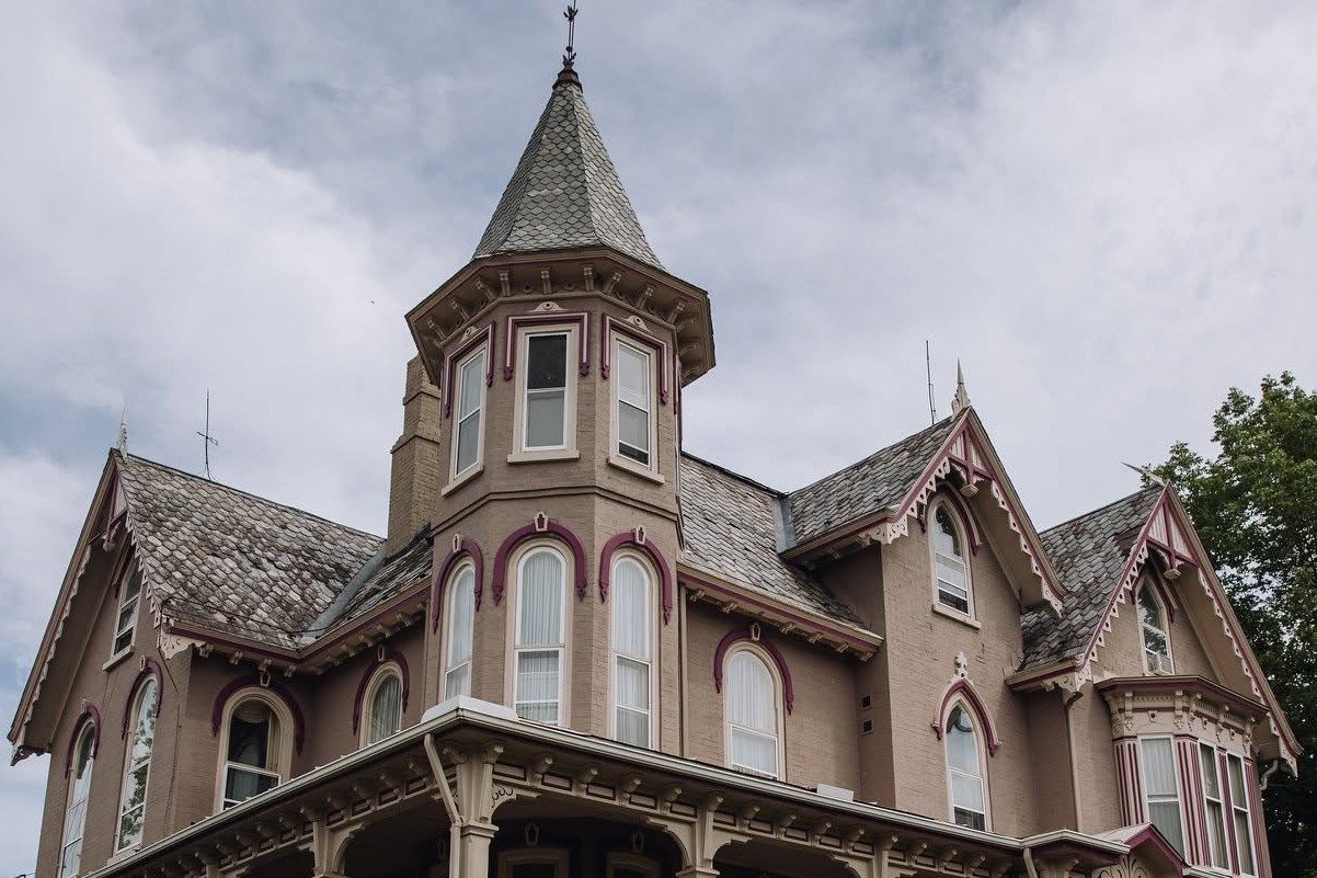 Victorian-style house with a turret, beige walls, and pink decorative trim under a cloudy sky.