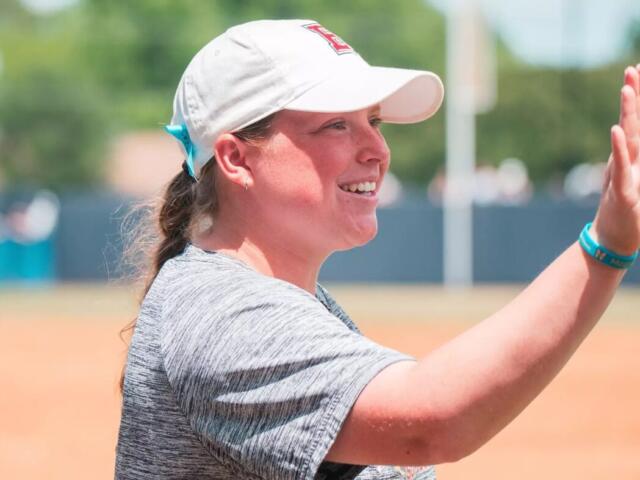 Smiling woman in a white cap and gray shirt gives a high-five on a baseball/softball field.