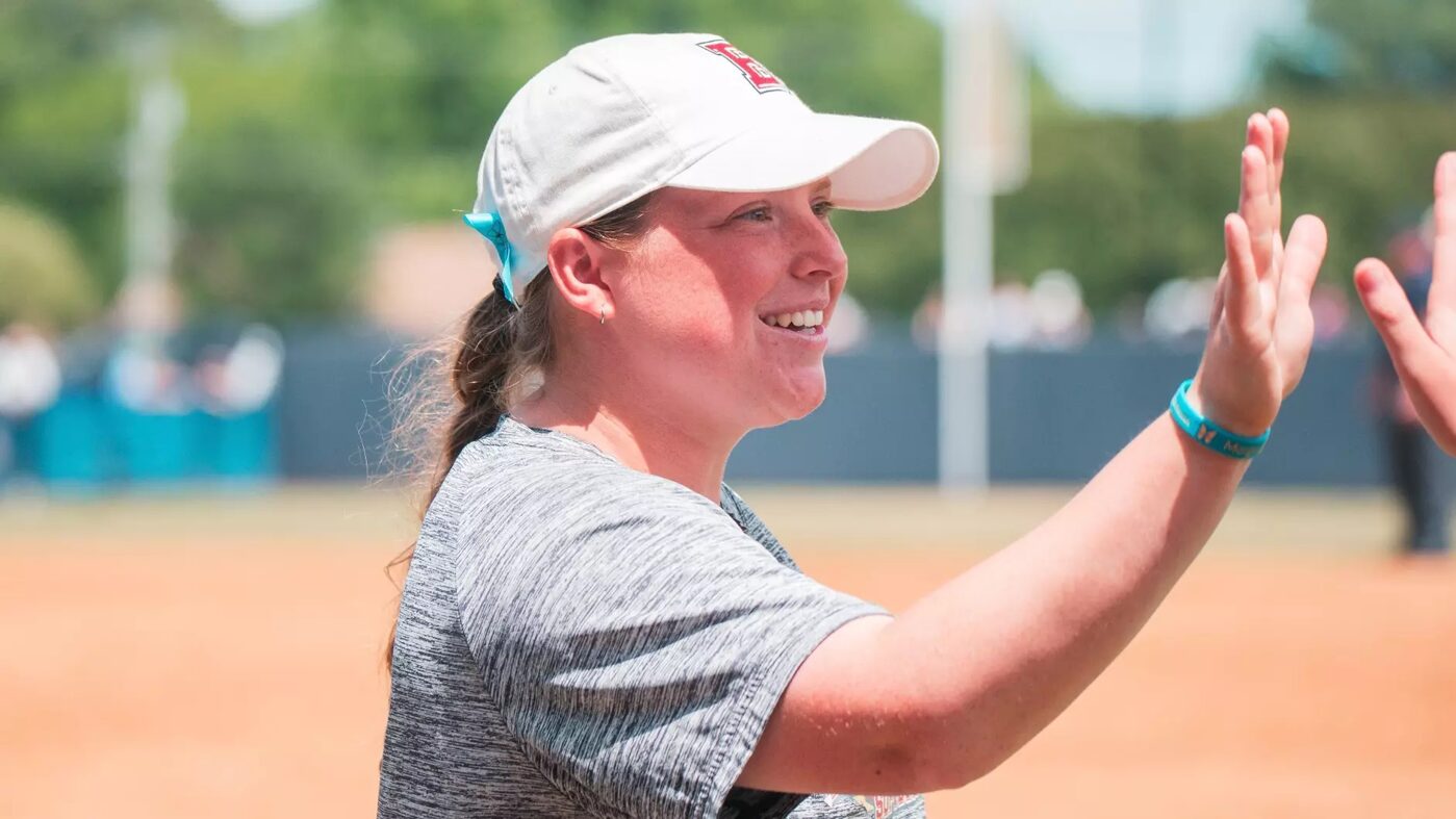 Smiling woman in a white cap and gray shirt gives a high-five on a baseball/softball field.