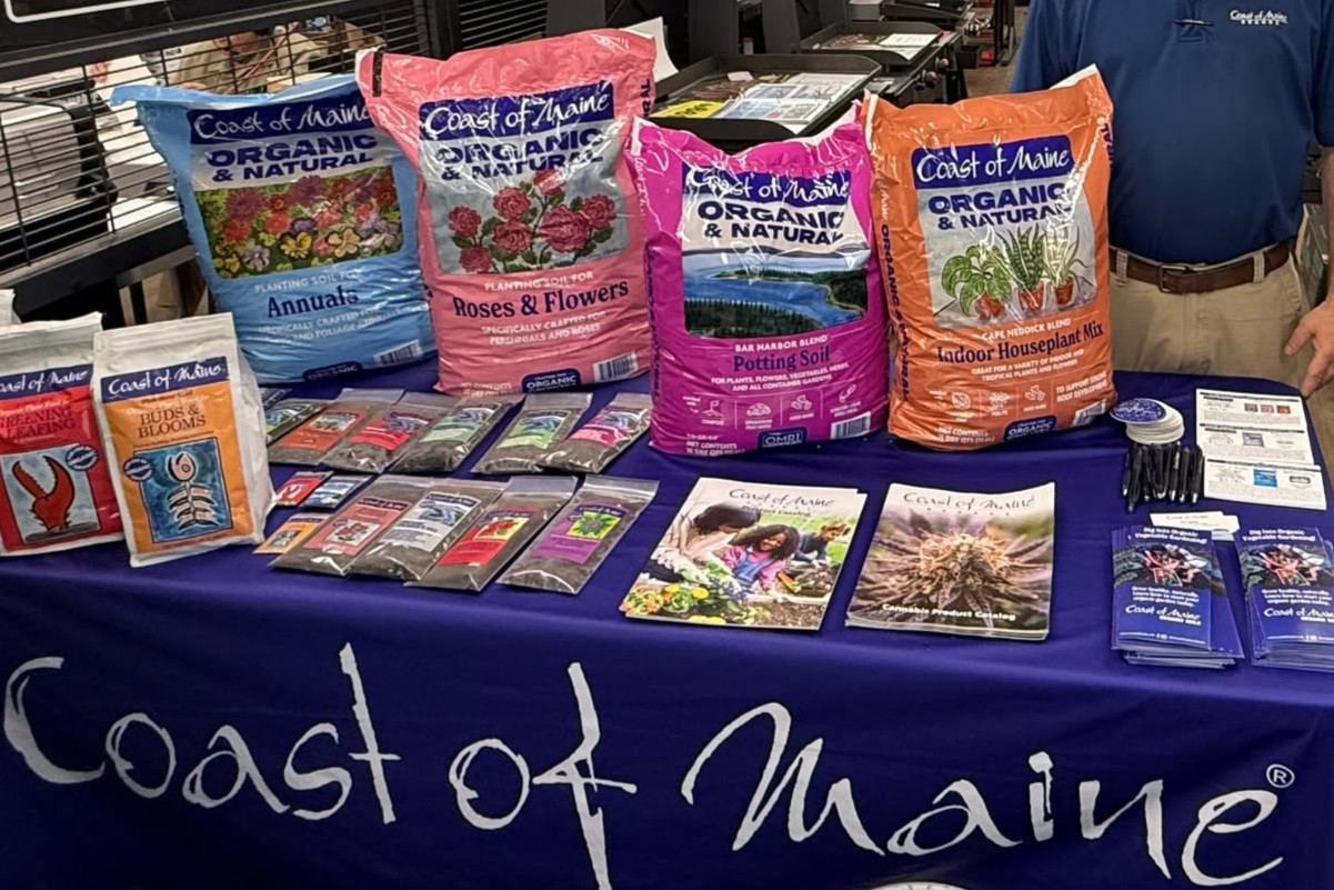 Coast of Maine gardening display with organic soil bags on a blue tablecloth, plus brochures and seed packets nearby.
