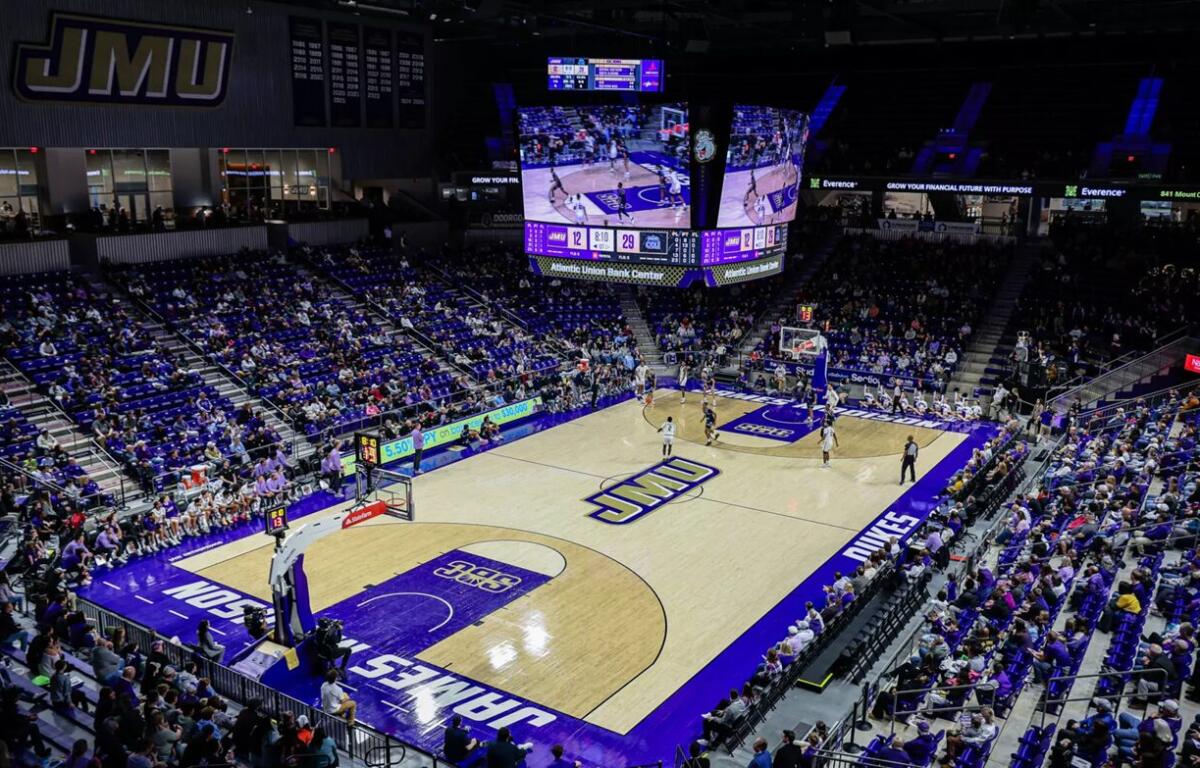 Wide shot of a college basketball game in a JMU arena, with a glowing scoreboard overhead and packed purple seats.