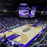 Wide shot of a college basketball game in a JMU arena, with a glowing scoreboard overhead and packed purple seats.