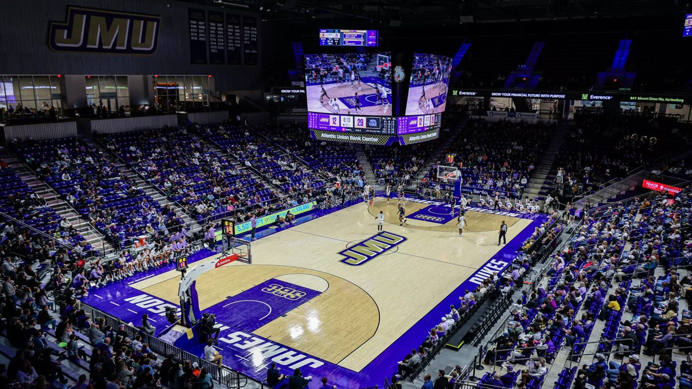 Wide shot of a college basketball game in a JMU arena, with a glowing scoreboard overhead and packed purple seats.