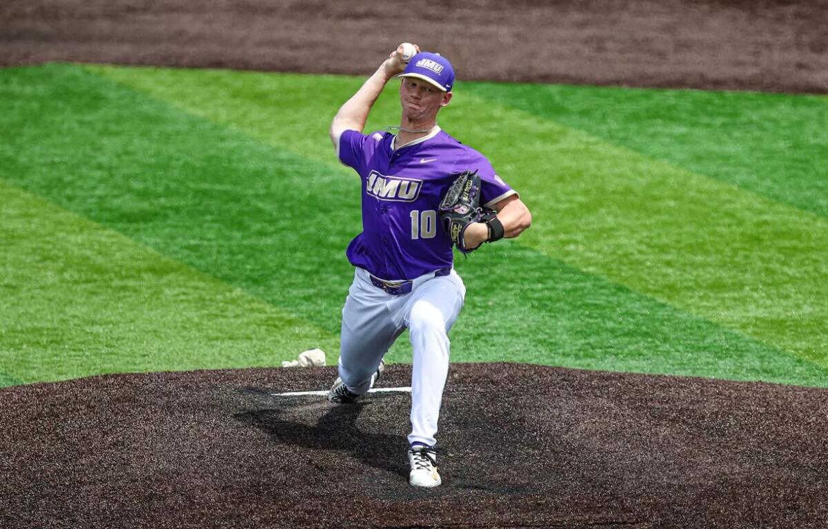 Baseball pitcher in a purple jersey winds up to throw from the pitcher's mound on a green baseball field.