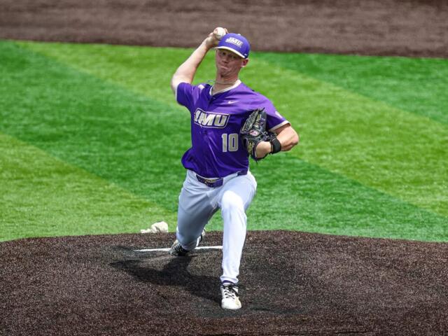 Baseball pitcher in a purple jersey winds up to throw from the pitcher's mound on a green baseball field.