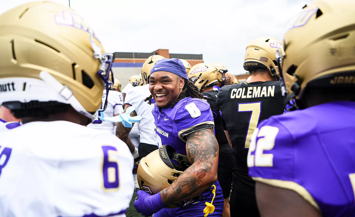 A smiling football player in a purple jersey stands among teammates in gold helmets on the sideline.