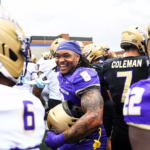 A smiling football player in a purple jersey stands among teammates in gold helmets on the sideline.