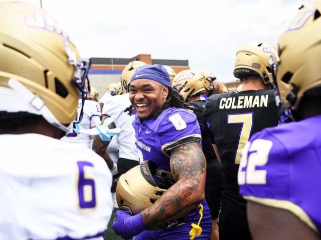 A smiling football player in a purple jersey stands among teammates in gold helmets on the sideline.