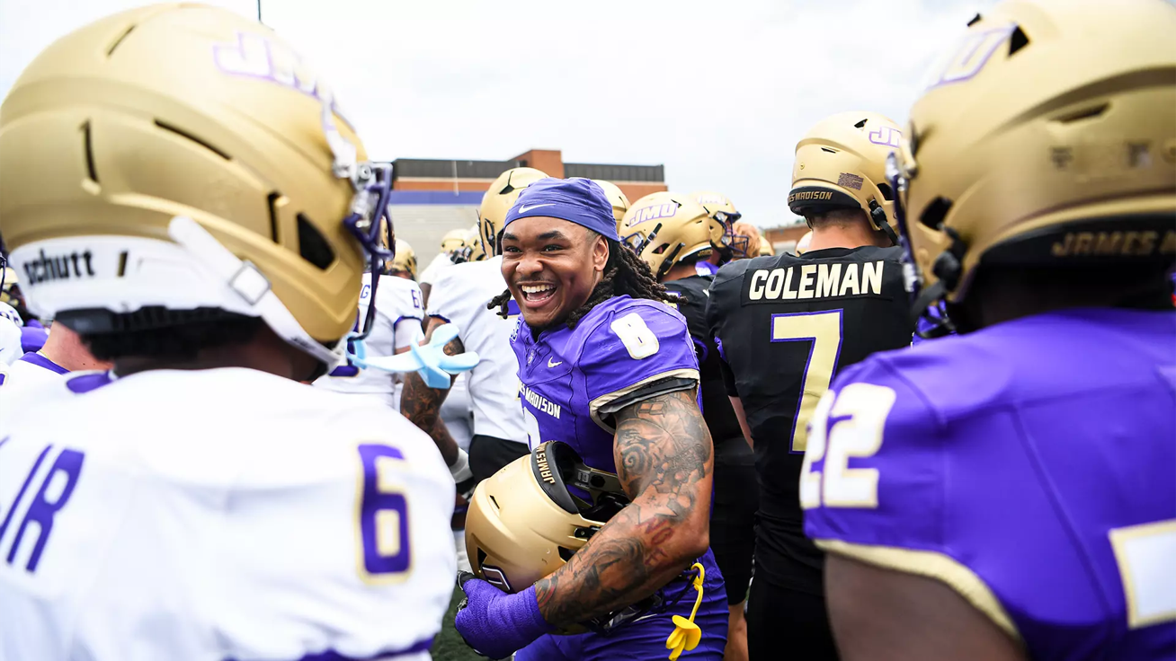 A smiling football player in a purple jersey stands among teammates in gold helmets on the sideline.