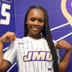 Smiling Black woman wearing a white pinstripe JMU jersey, posing in front of a purple mural wall with a mascot graphic.