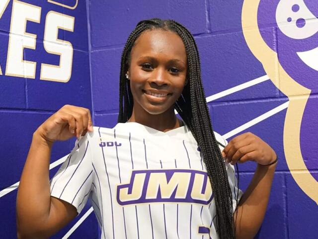 Smiling Black woman wearing a white pinstripe JMU jersey, posing in front of a purple mural wall with a mascot graphic.