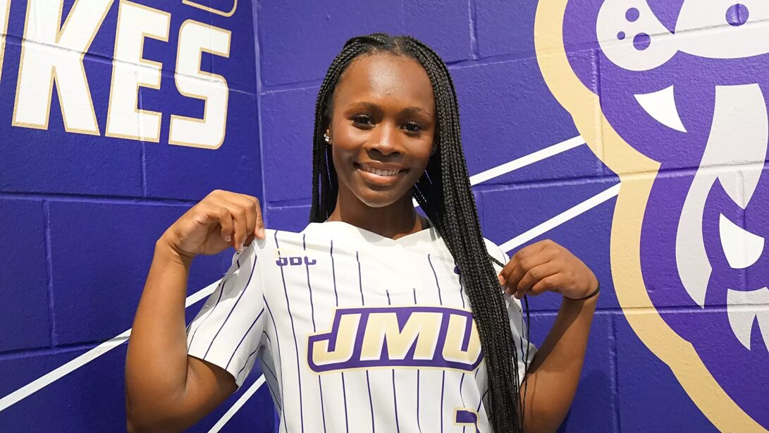 Smiling Black woman wearing a white pinstripe JMU jersey, posing in front of a purple mural wall with a mascot graphic.