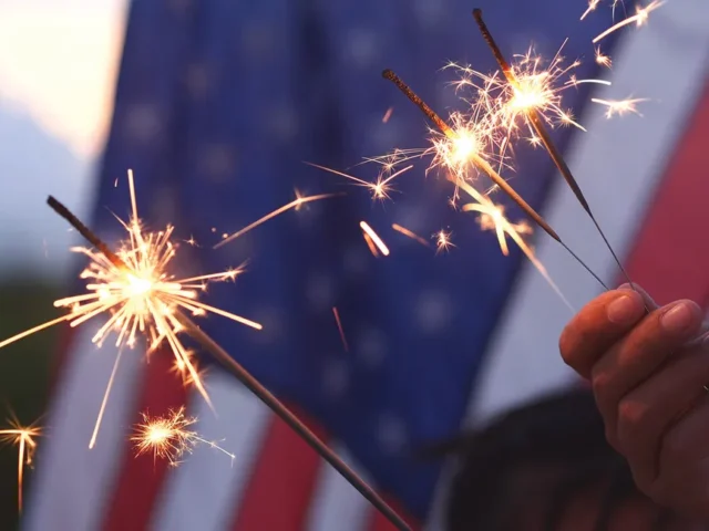 Close-up of a hand holding three lit sparklers with sparks flying, a blurred U.S. flag in the background.