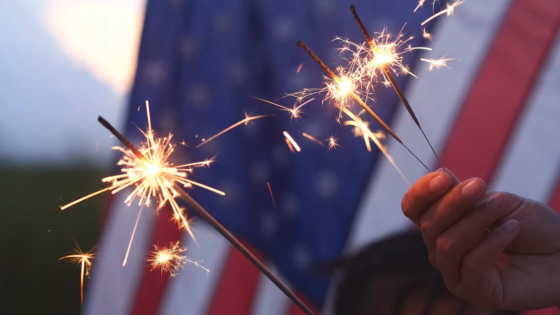 Close-up of a hand holding three lit sparklers with sparks flying, a blurred U.S. flag in the background.
