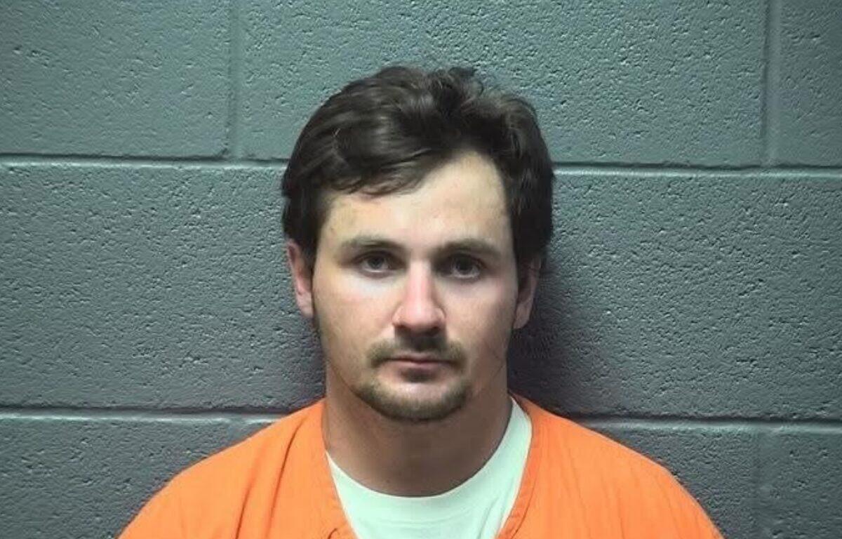 Front-facing mugshot of a man with short dark hair, wearing an orange jumpsuit, against a gray cinder block wall.