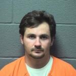 Front-facing mugshot of a man with short dark hair, wearing an orange jumpsuit, against a gray cinder block wall.