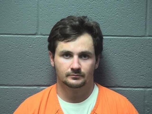 Front-facing mugshot of a man with short dark hair, wearing an orange jumpsuit, against a gray cinder block wall.