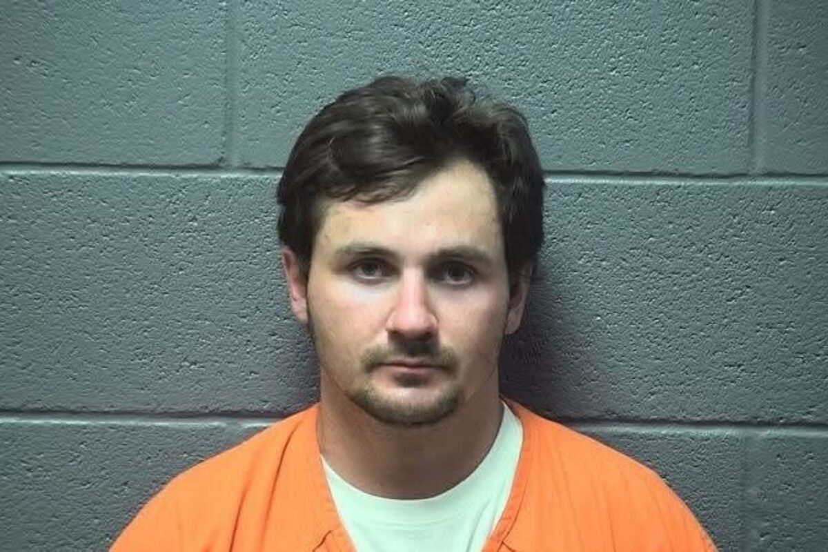 Front-facing mugshot of a man with short dark hair, wearing an orange jumpsuit, against a gray cinder block wall.
